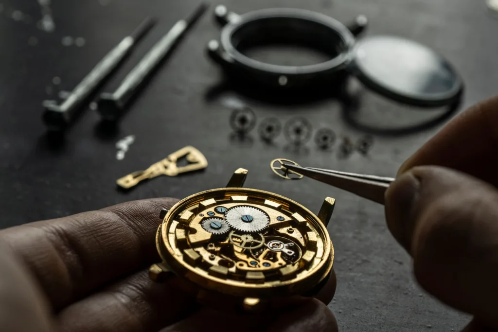 A picture of a mechanical watch undergoing repair. The watchmaker is holding a golden gear and is about to insert it into the watch.